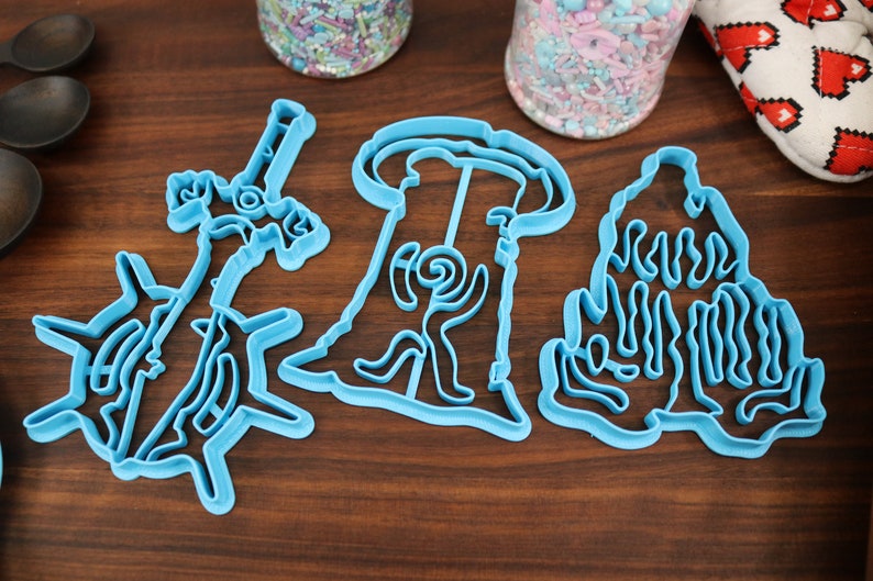 Puede incluir: Tres cortadores de galletas de pl&aacute;stico azul con forma de &aacute;rbol, casa y unicornio. Los cortadores est&aacute;n sobre una superficie de madera.