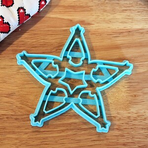 Pu&ograve; includere: Tagliapasta a forma di stella in plastica azzurra con intagli intricati, appoggiato su una superficie di legno. Il tagliapasta ha un design a stella a cinque punte con piccoli dettagli a stella all'interno di ogni punta.