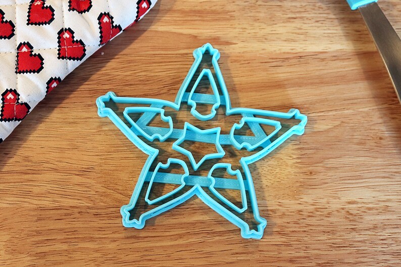 Pu&ograve; includere: Tagliapasta a forma di stella in plastica azzurra con intagli intricati, appoggiato su una superficie di legno. Il tagliapasta ha un design a stella a cinque punte con piccoli dettagli a stella all'interno di ogni punta.