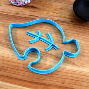 May include: A blue plastic cookie cutter shaped like a leaf with a smaller leaf design inside. The cutter is on a wooden surface.