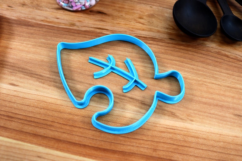 May include: A blue plastic cookie cutter shaped like a leaf with a smaller leaf design inside. The cutter is on a wooden surface.