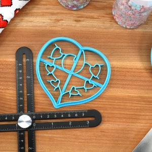 May include: A blue plastic heart-shaped cookie cutter with a design of smaller hearts inside. The cutter is on a wooden surface with a ruler and a blue spatula.