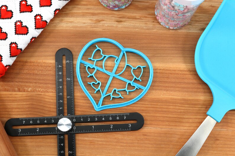 May include: A blue plastic heart-shaped cookie cutter with a design of smaller hearts inside. The cutter is on a wooden surface with a ruler and a blue spatula.