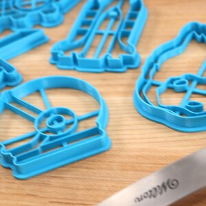 May include: A set of blue plastic cookie cutters shaped like various Star Wars characters, including Darth Vader, R2-D2, and a Stormtrooper. The cutters are on a wooden surface.