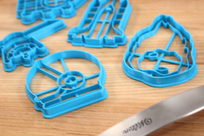 May include: A set of blue plastic cookie cutters shaped like various Star Wars characters, including Darth Vader, R2-D2, and a Stormtrooper. The cutters are on a wooden surface.