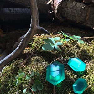 May include: Three teal green, translucent, geometrically shaped, resin gems on a bed of moss. The gems are reminiscent of a video game item.
