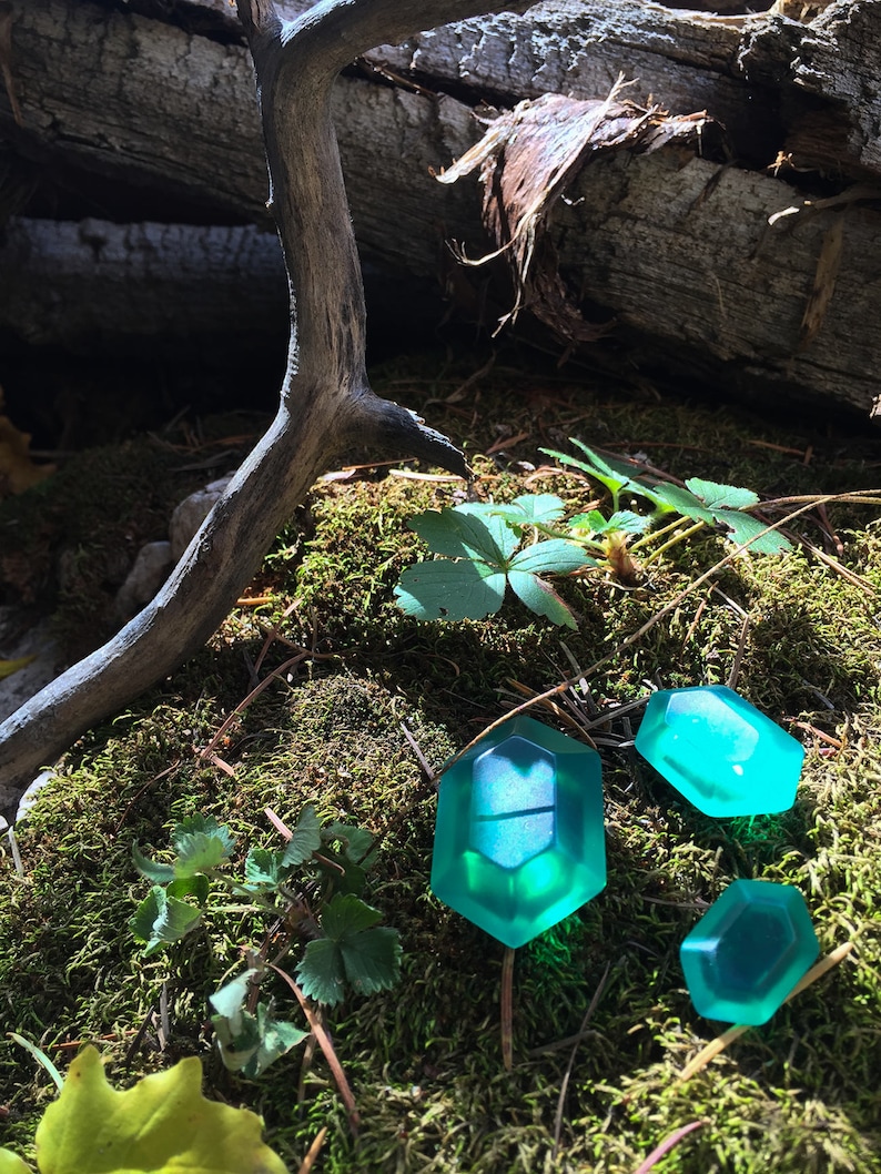 May include: Three teal green, translucent, geometrically shaped, resin gems on a bed of moss. The gems are reminiscent of a video game item.