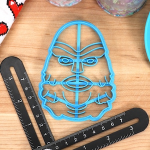 May include: A light blue plastic cookie cutter in the shape of a creature's face, with detailed facial features. A black ruler and a red and white heart-patterned cloth are nearby. A blue spatula is also visible.