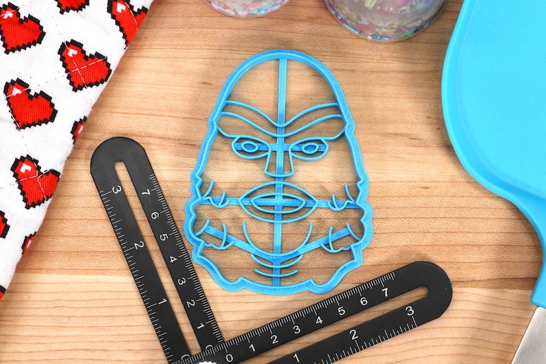 May include: A light blue plastic cookie cutter in the shape of a creature's face, with detailed facial features. A black ruler and a red and white heart-patterned cloth are nearby. A blue spatula is also visible.