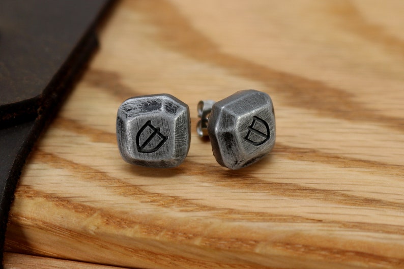 May include: A pair of silver stud earrings with a geometric design and a shield symbol engraved on each earring.