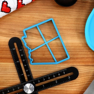 May include: A blue plastic cookie cutter shaped like a house with four windows. The cutter is on a wooden surface with a black ruler and a blue spatula.