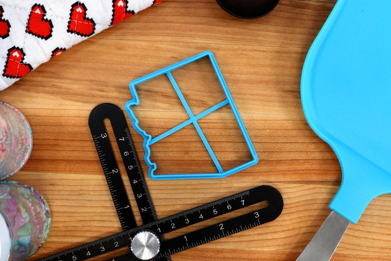 May include: A blue plastic cookie cutter shaped like a house with four windows. The cutter is on a wooden surface with a black ruler and a blue spatula.
