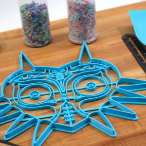 May include: A blue plastic cookie cutter shaped like the iconic Majora's Mask from the Legend of Zelda video game series. The cutter is on a wooden surface.