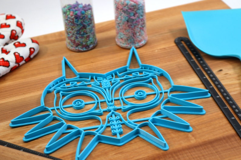 May include: A blue plastic cookie cutter shaped like the iconic Majora's Mask from the Legend of Zelda video game series. The cutter is on a wooden surface.