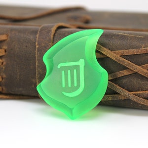 May include: A translucent green shield-shaped charm with a white symbol, in front of a brown leather journal with stitched detailing. The charm has a unique, stylized design, and the journal has a rustic appearance.