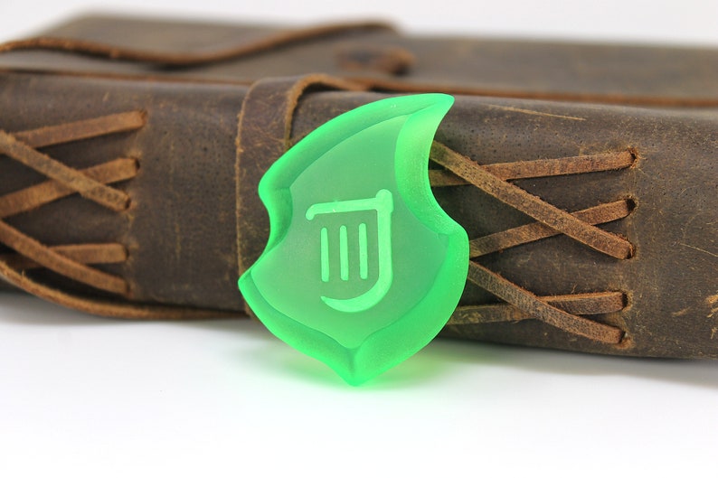 May include: A translucent green shield-shaped charm with a white symbol, in front of a brown leather journal with stitched detailing. The charm has a unique, stylized design, and the journal has a rustic appearance.