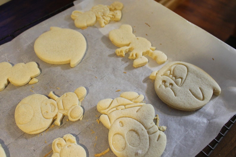 May include: A baking sheet lined with parchment paper holds nine unbaked sugar cookies. The cookies are in various shapes, including a circle, a snowman, a cartoon character, and a heart. The cookies are a light golden brown color.