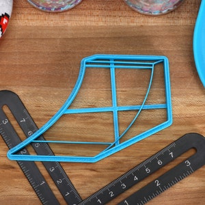May include: A blue plastic cookie cutter shaped like a shoe. The cutter is on a wooden surface with black rulers.