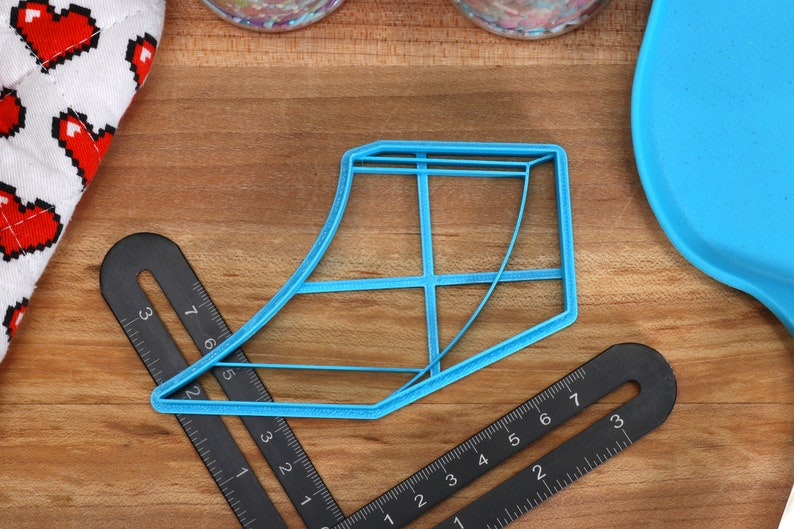 May include: A blue plastic cookie cutter shaped like a shoe. The cutter is on a wooden surface with black rulers.