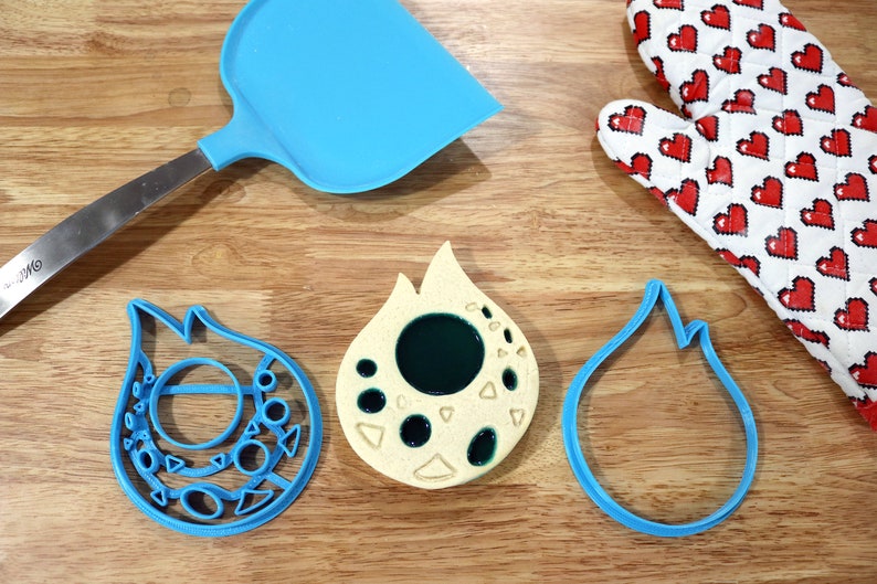 May include: Three blue cookie cutters shaped like flames, one with a circle and smaller shapes inside, and one with a filled-in green circle inside. A yellow cookie with a green circle inside and smaller shapes around it.
