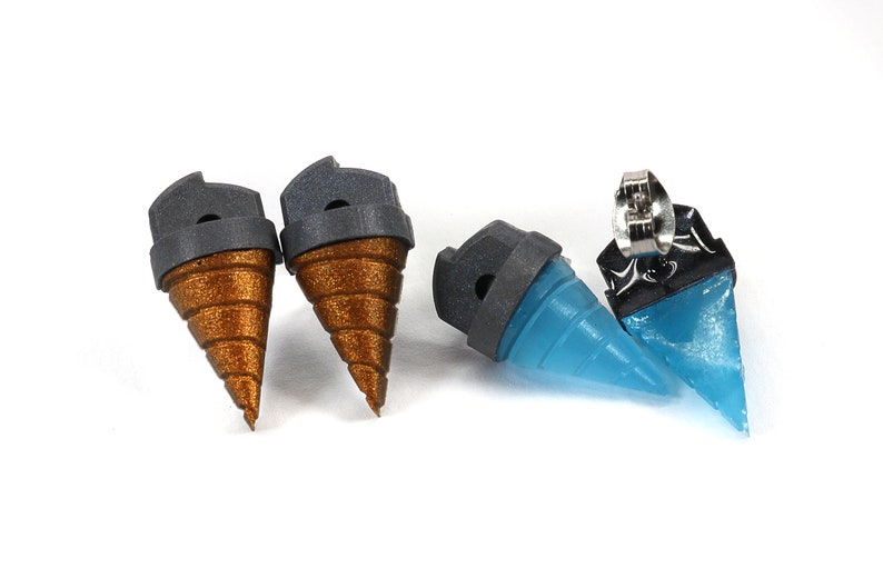 May include: A set of four earrings. Two are a metallic bronze color with a gray top, and two are a translucent blue with a black top. The earrings are shaped like a drill bit.