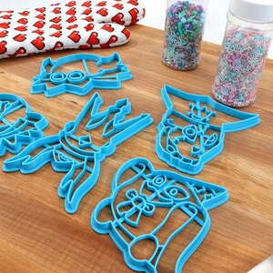 May include: A collection of blue plastic cookie cutters in various cartoon character shapes, arranged on a wooden surface. A red and white heart-patterned oven mitt and two jars of sprinkles are also visible. A blue spatula is in the background.