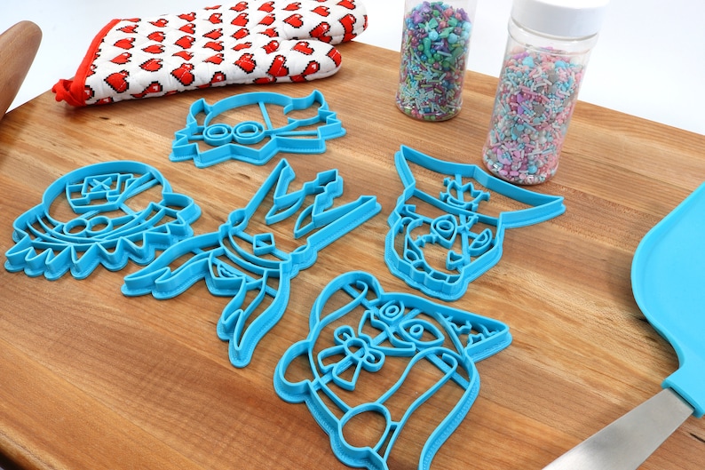 May include: A collection of blue plastic cookie cutters in various cartoon character shapes, arranged on a wooden surface. A red and white heart-patterned oven mitt and two jars of sprinkles are also visible. A blue spatula is in the background.