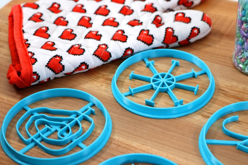 May include: Four blue plastic cookie cutters on a wooden surface. The cutters are shaped like a snowflake, a swirl, a hand, and a pair of scissors.