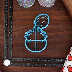 May include: A blue plastic cookie cutter shaped like a cartoon monster with a single eye and a large, round head. The monster has a spiky hairdo and is holding a balloon.
