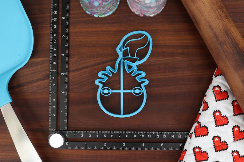 May include: A blue plastic cookie cutter shaped like a cartoon monster with a single eye and a large, round head. The monster has a spiky hairdo and is holding a balloon.