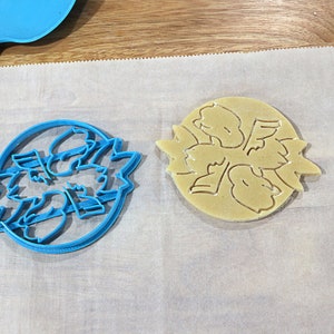 May include: A blue plastic cookie cutter and a light brown cookie on a white parchment paper surface. The cookie cutter and cookie are shaped like a flower with five petals.