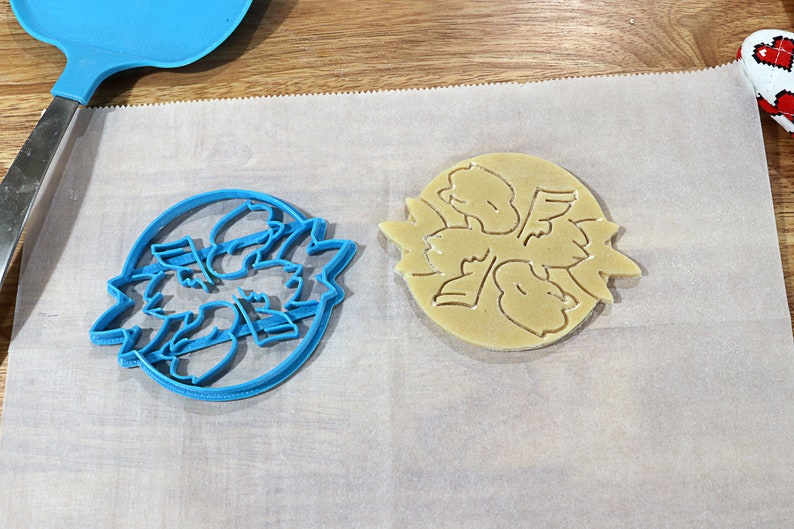 May include: A blue plastic cookie cutter and a light brown cookie on a white parchment paper surface. The cookie cutter and cookie are shaped like a flower with five petals.