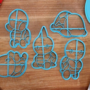May include: Five blue plastic cookie cutters shaped like cartoon turtles. The cutters have a grid pattern on them. The cutters are on a wooden surface.
