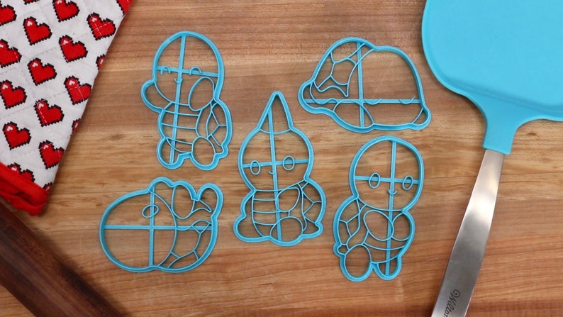 May include: Five blue plastic cookie cutters shaped like cartoon turtles. The cutters have a grid pattern on them. The cutters are on a wooden surface.