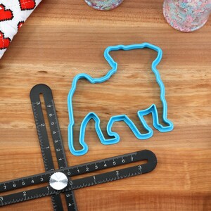May include: Blue plastic cookie cutter shaped like a pug dog on a wooden surface with a ruler and a red and white heart patterned fabric.