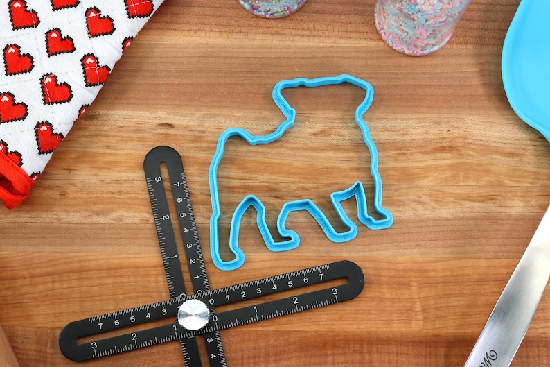 May include: Blue plastic cookie cutter shaped like a pug dog on a wooden surface with a ruler and a red and white heart patterned fabric.