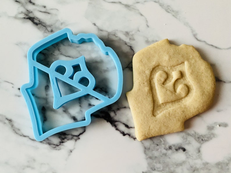 May include: A blue plastic cookie cutter shaped like a heart with the number 25 inside. A cookie is shown next to the cutter with the same design imprinted on it.