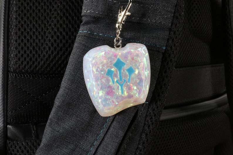 K&ouml;nnte beinhalten: Ein wei&szlig;er, schimmernder Schl&uuml;sselanh&auml;nger mit einem blauen Dreizack-Symbol. Der Schl&uuml;sselanh&auml;nger ist an einem schwarzen Rucksack befestigt.