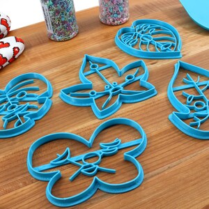 May include: Five blue plastic cookie cutters shaped like leaves. The cutters are on a wooden surface.