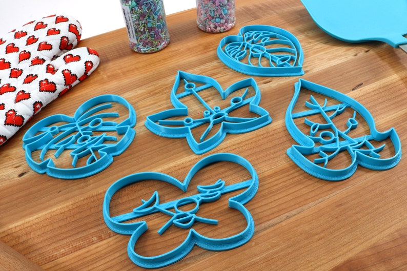 May include: Five blue plastic cookie cutters shaped like leaves. The cutters are on a wooden surface.