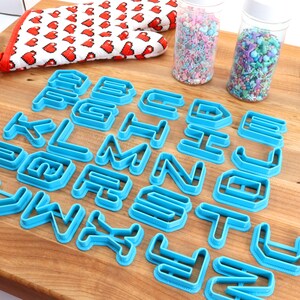 May include: A set of blue plastic alphabet cookie cutters on a wooden surface. The cutters are arranged in alphabetical order.