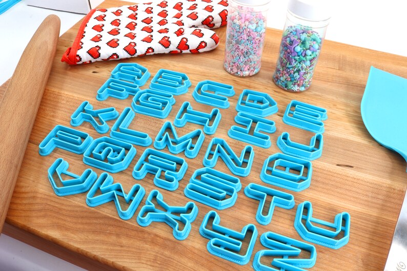 May include: A set of blue plastic alphabet cookie cutters on a wooden surface. The cutters are arranged in alphabetical order.