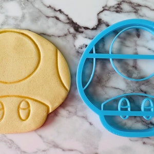May include: A blue plastic cookie cutter shaped like a mushroom. Next to it is a light brown cookie that has been cut out using the cookie cutter.