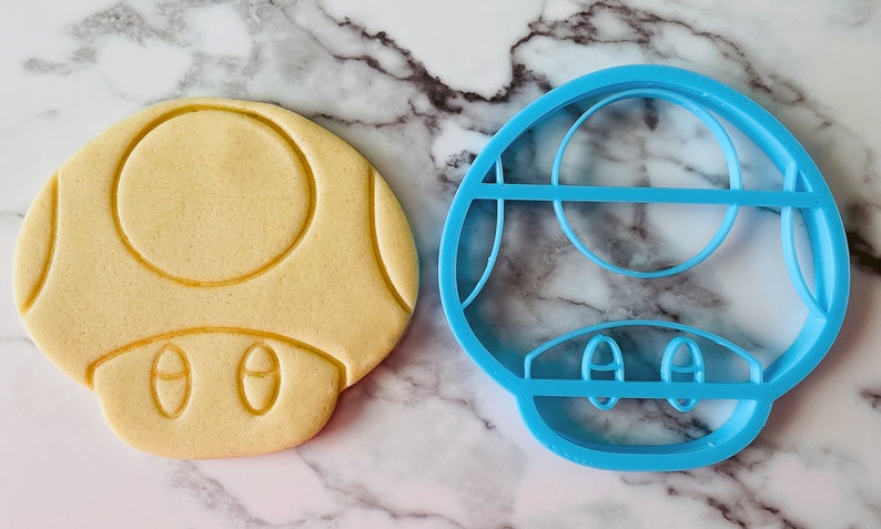May include: A blue plastic cookie cutter shaped like a mushroom. Next to it is a light brown cookie that has been cut out using the cookie cutter.