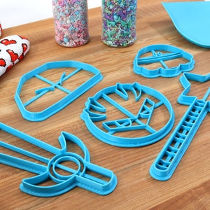 May include: A set of blue plastic cookie cutters shaped like a sword, a star, a shield, a crown, and a flower. The cutters are on a wooden surface.