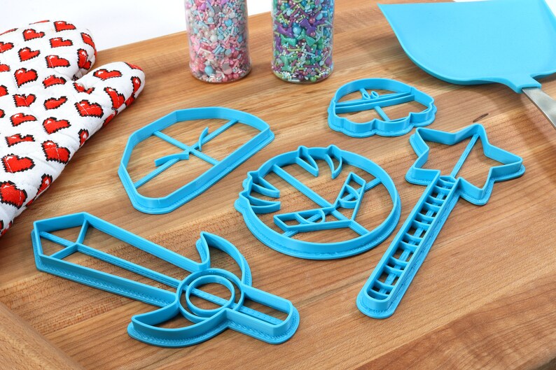 May include: A set of blue plastic cookie cutters shaped like a sword, a star, a shield, a crown, and a flower. The cutters are on a wooden surface.