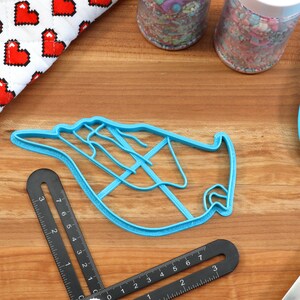 May include: A light blue plastic cookie cutter shaped like a hand with fingers extended. The cutter is on a wooden surface with other baking supplies.