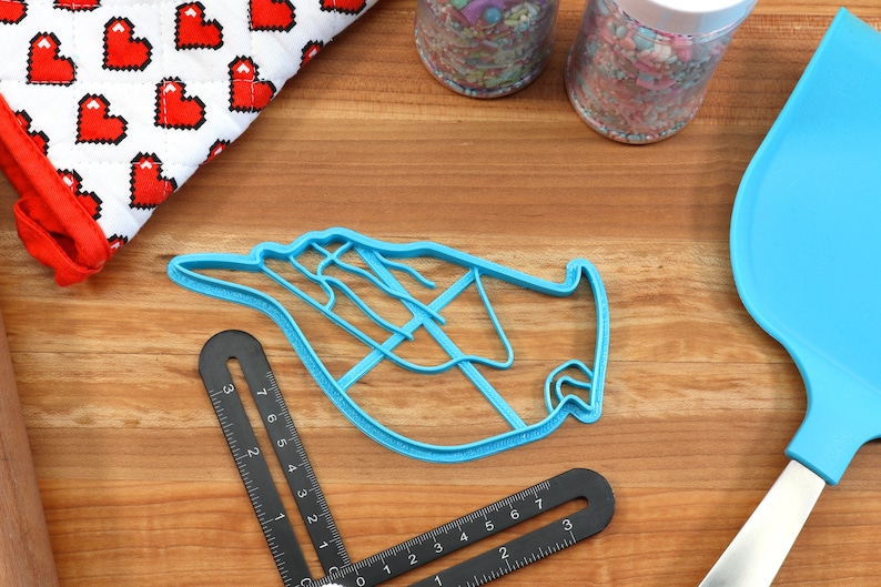 May include: A light blue plastic cookie cutter shaped like a hand with fingers extended. The cutter is on a wooden surface with other baking supplies.