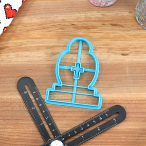 May include: Blue plastic cookie cutter shaped like a tombstone with a cross on the front.