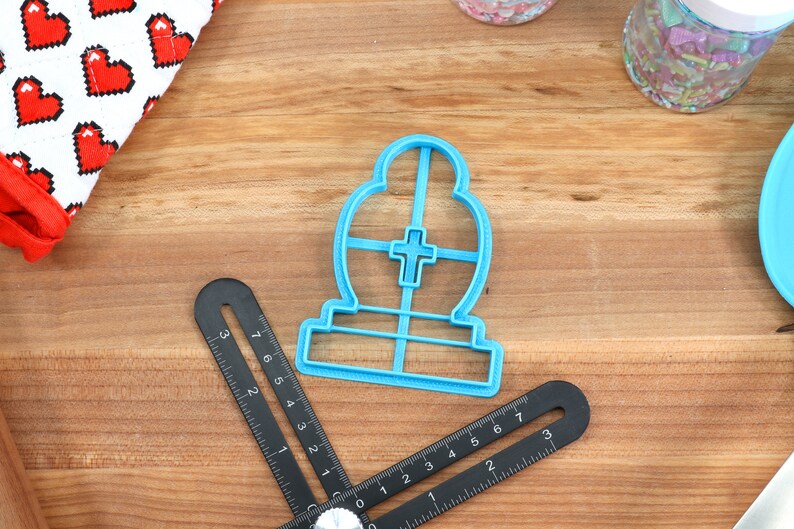May include: Blue plastic cookie cutter shaped like a tombstone with a cross on the front.